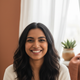 A smiling South Asian woman in her late 20s, headshot portrait with a warm terracotta and white background.